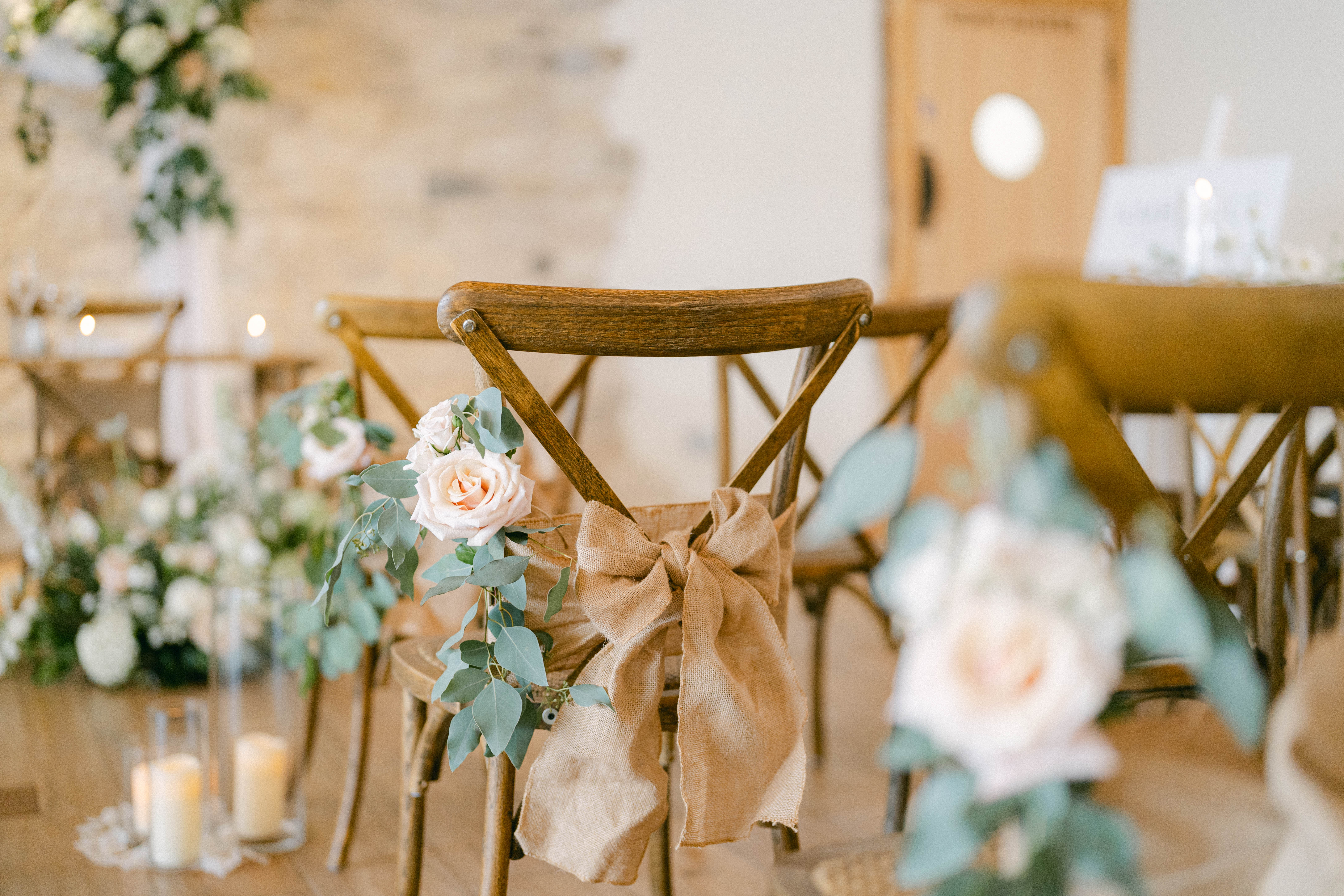 Wooden chairs with burlap bows and roses, set for a wedding. Candles and flowers decorate the aisle in a softly lit room. - Wild Thyme & Honey Hotel in the Cotswolds