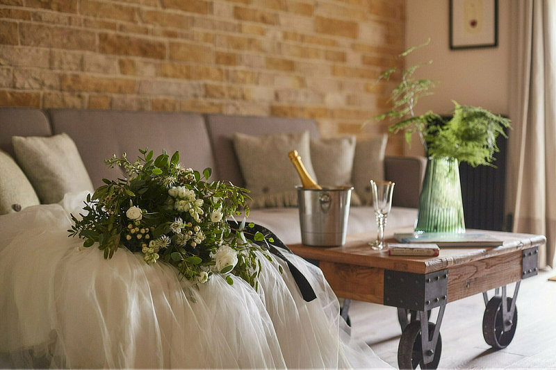 A cozy living room with a bouquet, champagne, and glasses on a wooden table. Neutral-toned decor and brick wall backdrop. - Wild Thyme & Honey Hotel in the Cotswolds