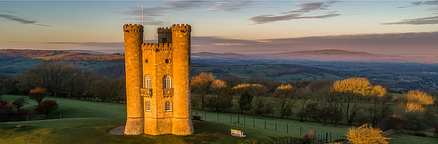A tall, historic tower on a hilltop during sunset, surrounded by fields and trees, with distant rolling hills under a colorful sky. - Wild Thyme & Honey Hotel in the Cotswolds