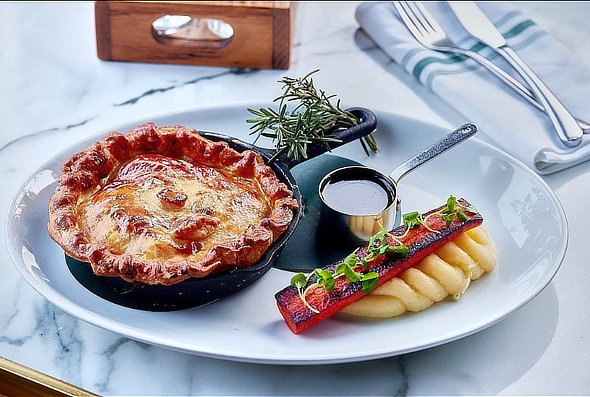 A gourmet pie in a black dish with a side of mashed potatoes, a strip of grilled vegetable, sauce, and rosemary garnish on a white plate. - Wild Thyme & Honey Hotel in the Cotswolds