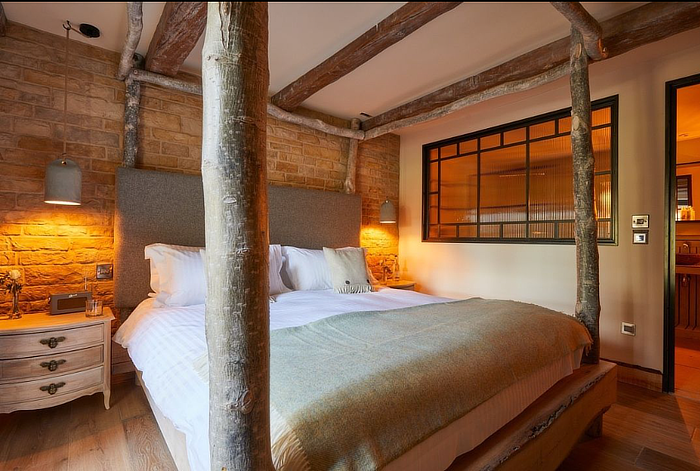 Cozy bedroom with a rustic wooden canopy bed, exposed brick wall, warm lighting, and a wooden nightstand. - Wild Thyme & Honey Hotel in the Cotswolds