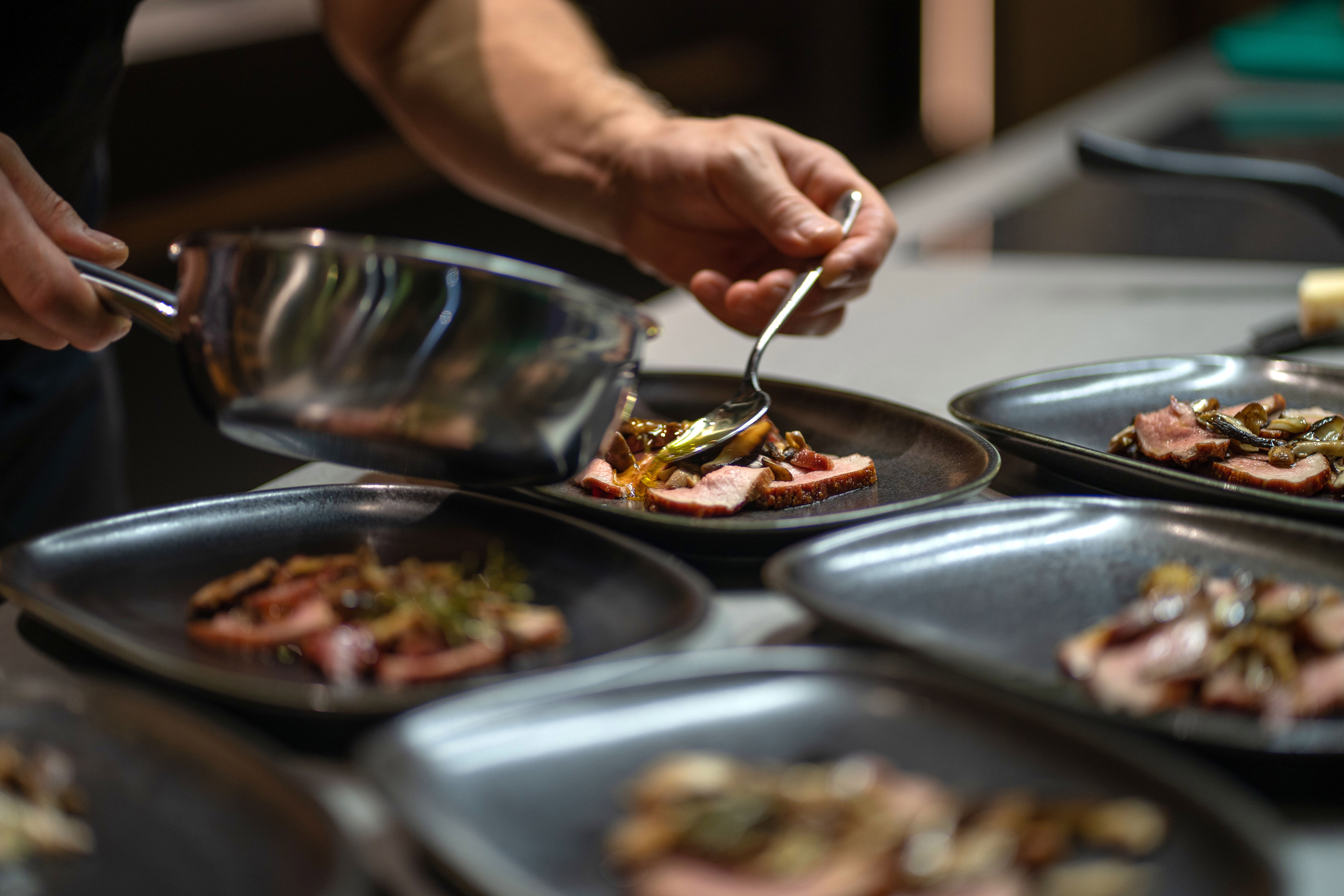 A chefs hands skillfully plating meat and vegetables onto several black dishes in a kitchen. A saucepan is held in one hand while a spoon is used to arrange the food, suggesting a professional culinary setting. - Wild Thyme & Honey Hotel in the Cotswolds