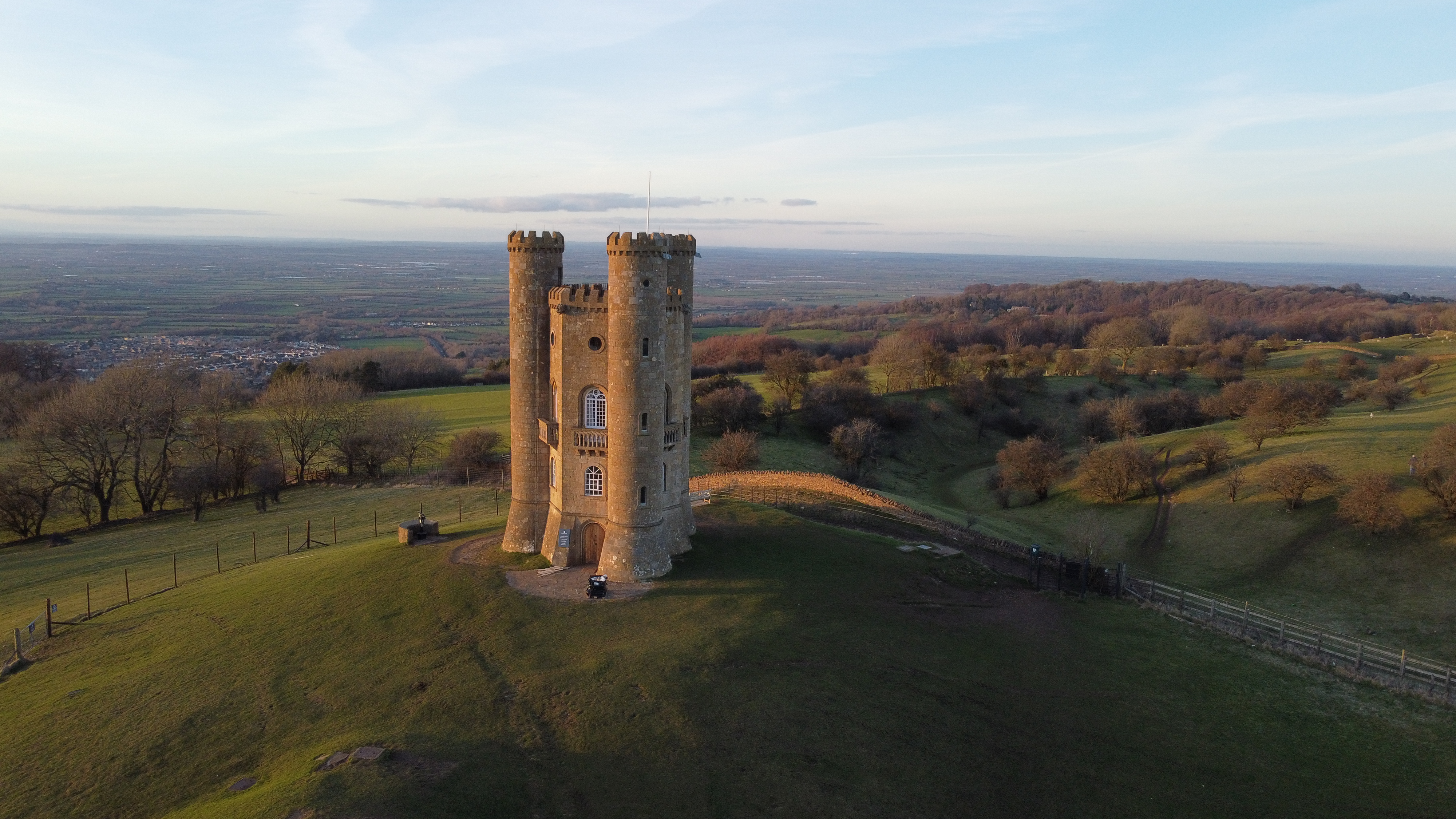 Broadway Tower on a grassy hilltop during sunset, surrounded by fields and trees with a distant view of the landscape below. - Wild Thyme & Honey Hotel in the Cotswolds