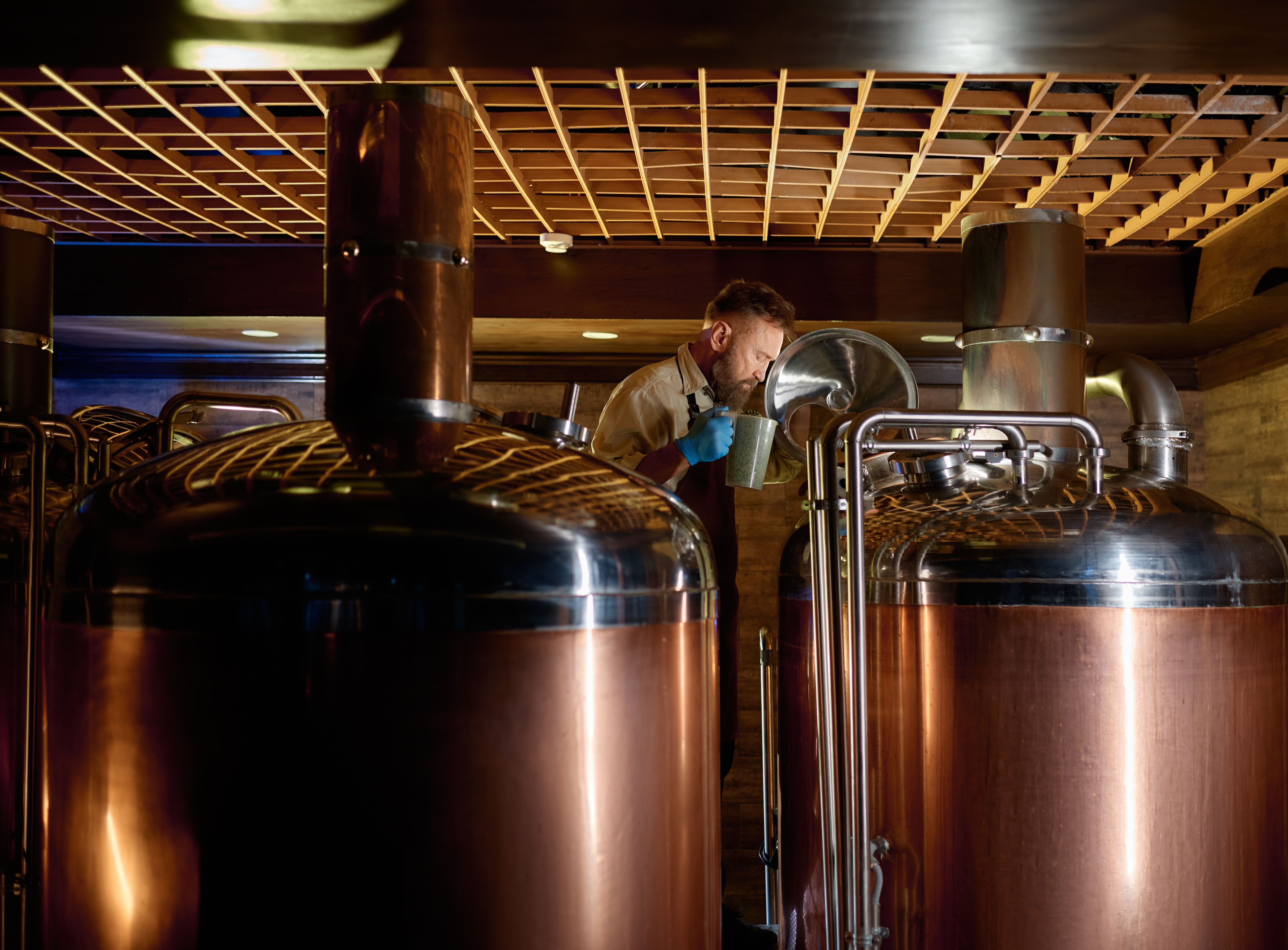 A person carefully inspecting a liquid sample from a large copper brewing tank in a dimly lit brewery setting. - Wild Thyme & Honey Hotel in the Cotswolds