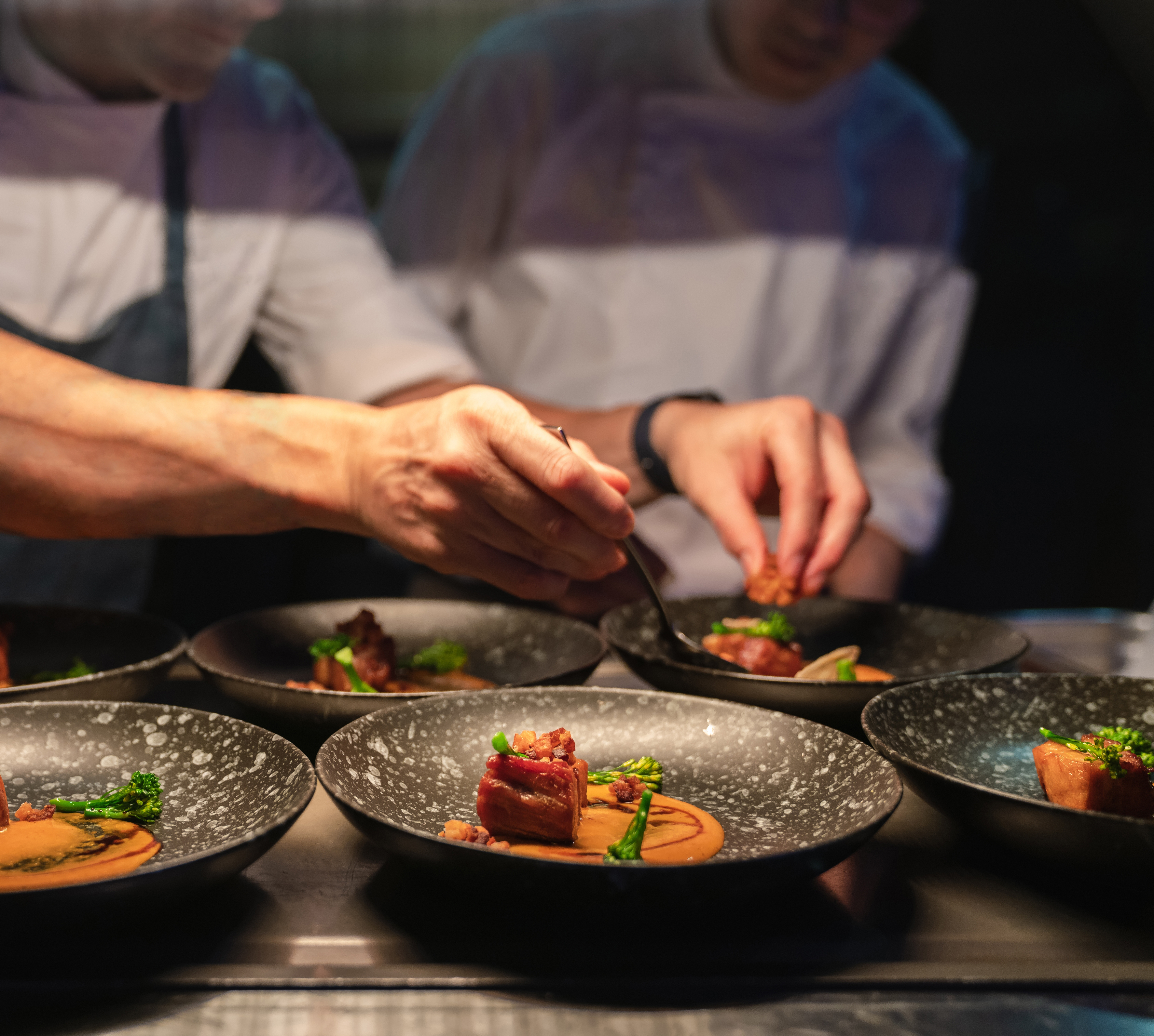 Chefs delicately plating gourmet dishes with meat, sauce, and vegetables on dark plates in a restaurant kitchen. - Wild Thyme & Honey Hotel in the Cotswolds