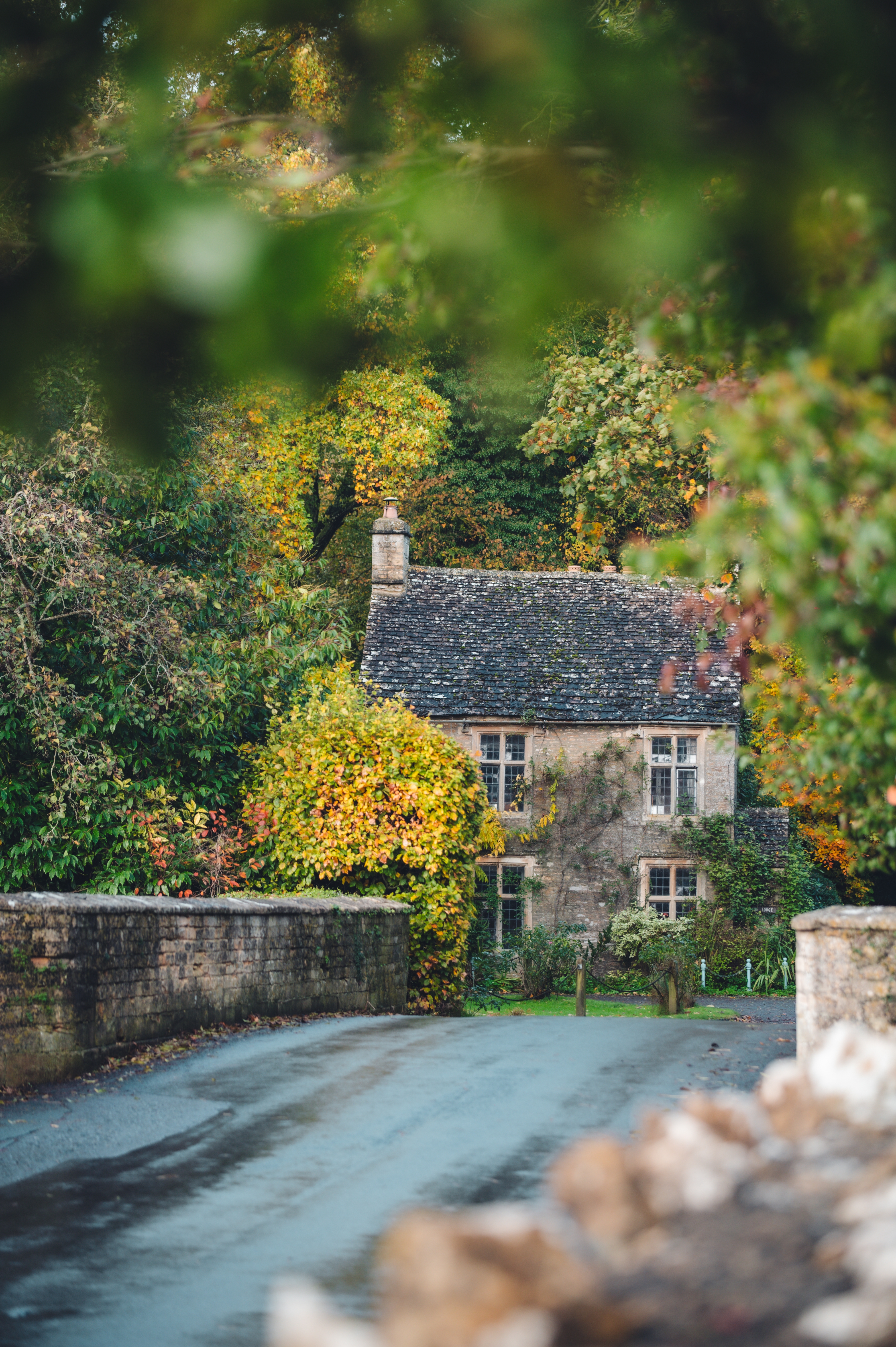 Charming stone cottage surrounded by autumn trees, viewed from a country road with a stone wall in the foreground. - Wild Thyme & Honey Hotel in the Cotswolds