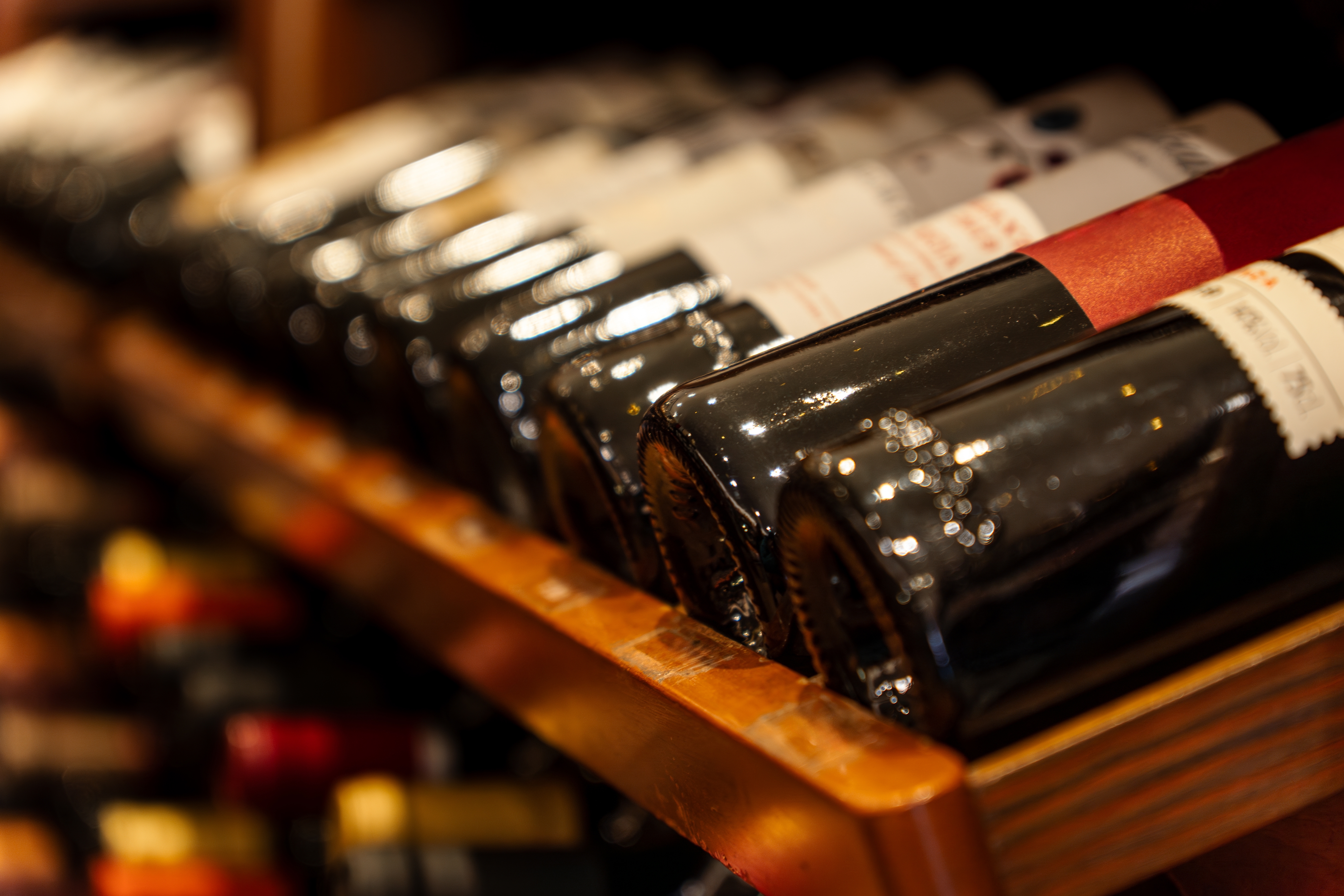 Close-up of wine bottles lined horizontally on a wooden shelf, with some labels visible and light reflecting off the glass. - Wild Thyme & Honey Hotel in the Cotswolds