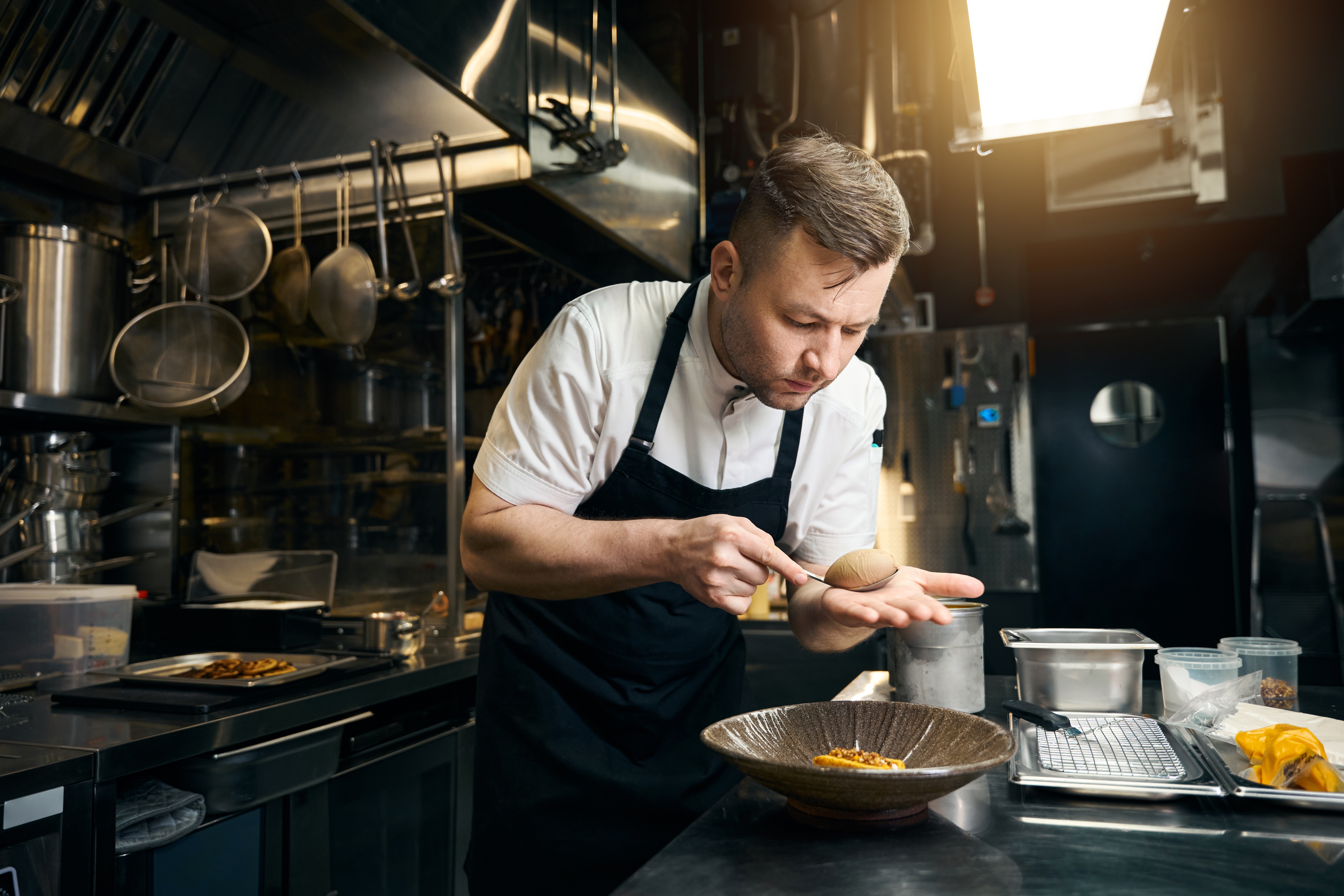 Chef in a professional kitchen carefully plating food with a spoon, wearing a black apron and focused on presentation. - Wild Thyme & Honey Hotel in the Cotswolds