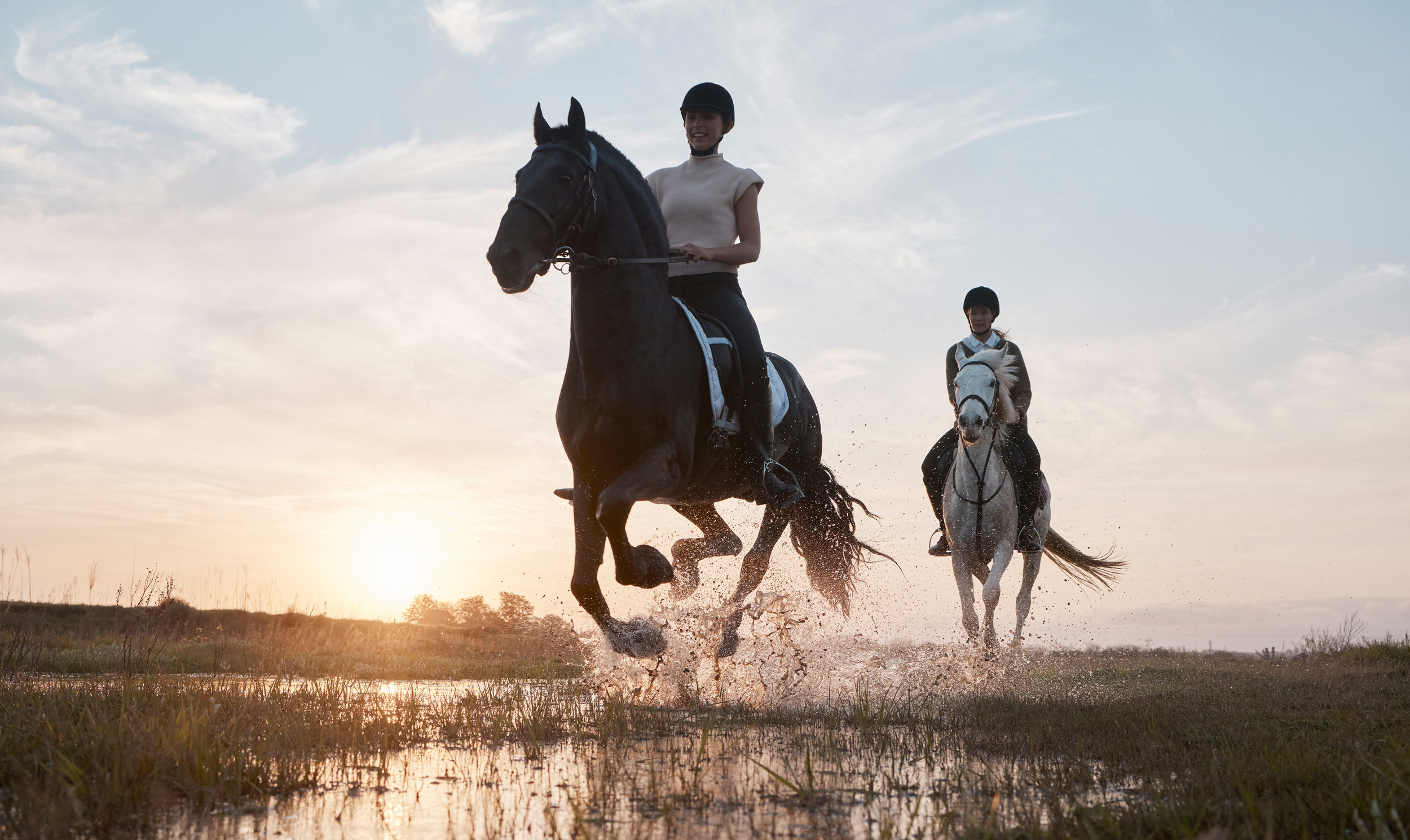 Two people riding horses through a wet field at sunset. The rider in the foreground is on a black horse, while the rider behind is on a white horse. Water splashes around as the horses gallop, with a colorful sky in the background. - Wild Thyme & Honey Hotel in the Cotswolds