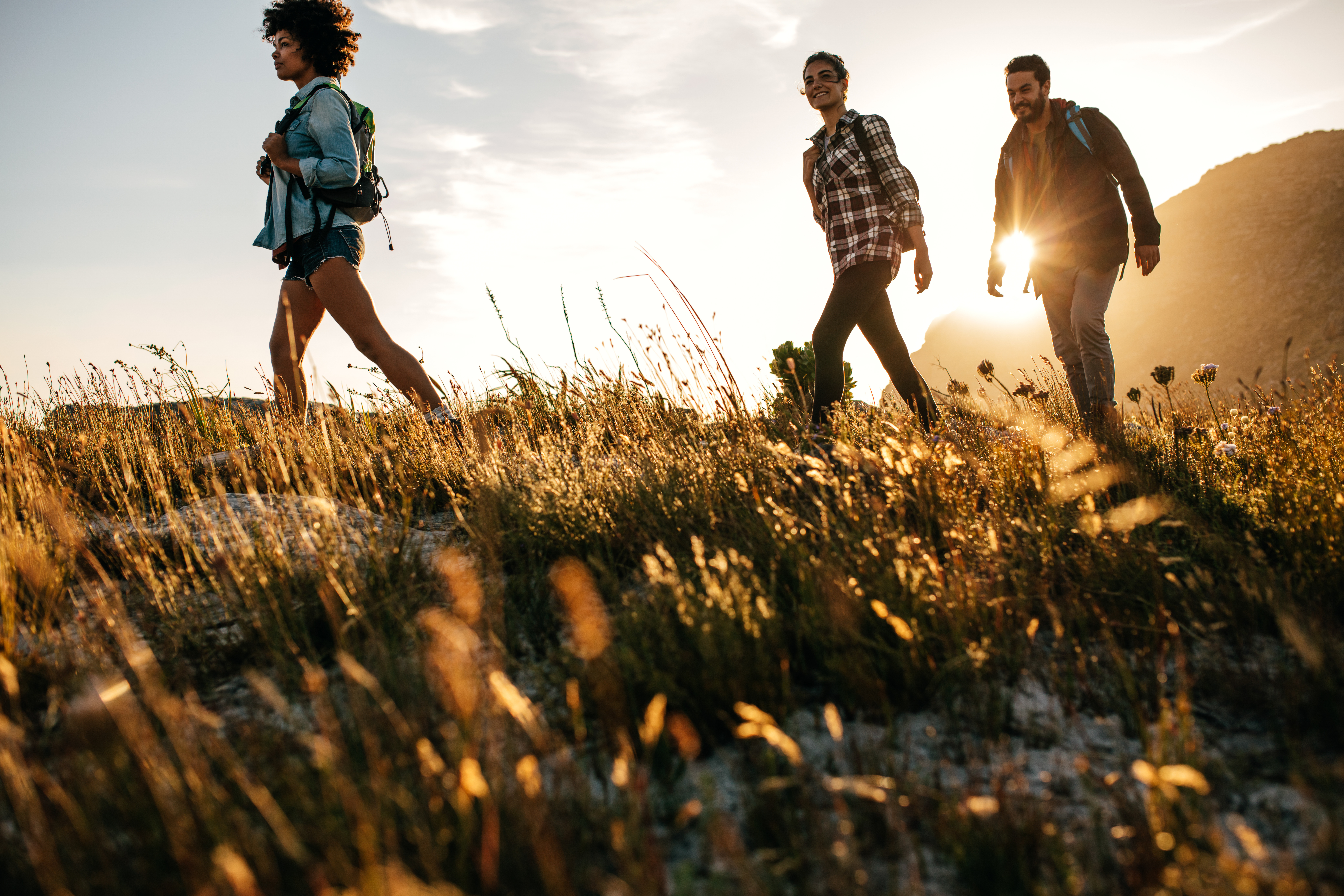 Three people hiking in a grassy field at sunset, with mountains in the background. Sunlight creates a warm glow around them. - Wild Thyme & Honey Hotel in the Cotswolds